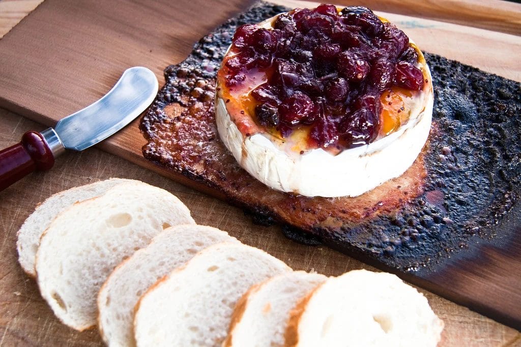 cylindrical brie on a wooden plaque with a chutney in top and some slices of bread on the side, placed next to a cheese knife.